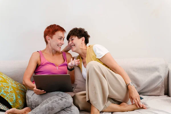 熟女レズビアンカップル使用してラップトップの間に座っています上のソファ一緒にホーム — ストック写真