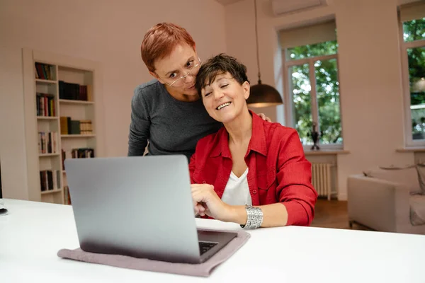 Dojrzałe Lesbijskie Para Uśmiecha Się Przytula Podczas Korzystania Laptopa Domu — Zdjęcie stockowe