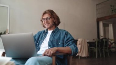 Gözlüklü, gülümseyen sarışın adam kafede laptopla konuşuyor.