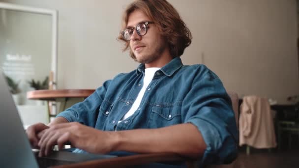 笑顔ブロンド男で眼鏡タイプのラップトップでカフェ — ストック動画
