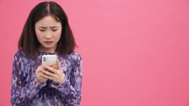 Memnun olmayan Asyalı kadın pembe stüdyoda cep telefonuyla yazı yazıyor.