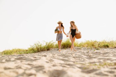 Genç iki kadın hasır şapkalar takıp kumsalda sepetle yürüyor.