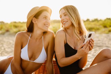 Mayolu iki genç kadın sahilde gülümsüyor ve cep telefonu kullanıyor.