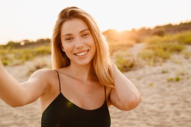 Kumsalda selfie çekerken gülümseyen genç sarışın kadın.