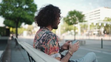 Kıvırcık saçlı gözlüklü adam bankta otururken cep telefonuyla mesaj atıyor.