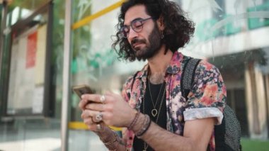 Kıvırcık saçlı, gözlüklü bir adam otobüs durağında telefonla mesajlaşıyor.