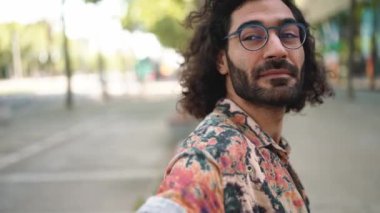 Kıvırcık saçlı, gözlüklü, pozitif sakallı adam bankta otururken etrafına bakıyor.