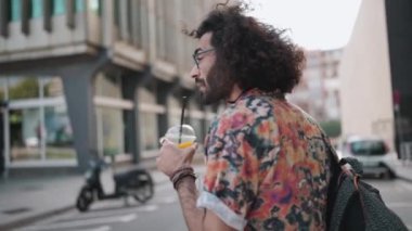 Gözlüklü, kıvırcık saçlı, meyve suyu içip dışarıda yürüyen adam.