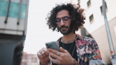 Dalgın kıvırcık saçlı, gözlüklü adam telefona ve dışarı bakıyor.