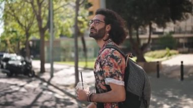 Ciddi kıvırcık saçlı, gözlüklü, dışarıda meyve suyuyla yürüyen sakallı bir adam.