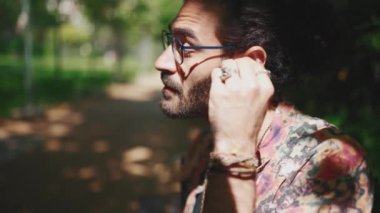 Ciddi kıvırcık saçlı, gözlüklü, dışarıda bankta kulaklık takan sakallı adam.