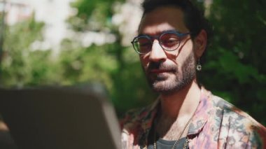Kendine güvenen, kıvırcık saçlı, gözlüklü, tablete bakan, açık hava bankında oturan.