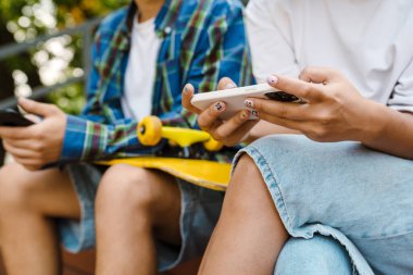 Dışarıda kaykay parkında zaman geçirirken gülümseyen ve cep telefonu kullanan iki çok kültürlü genç.