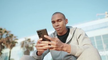 Sabah dışarıda otururken telefonla mesajlaşan kel bir sporcu.