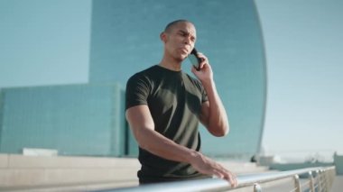 Siyah tişört giyen, gülen kel sporcu sabahları dışarıda cep telefonuyla konuşuyor.