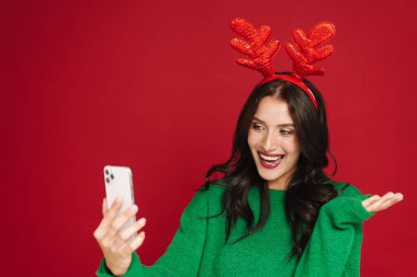 Geyik boynuzları takan genç bir kadın kırmızı duvarda izole edilmiş bir cep telefonu kullanıyor.