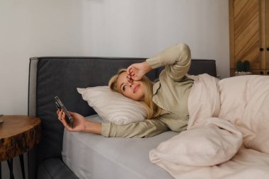 Evde uyuduktan sonra yatakta uzanan genç sarışın kadın cep telefonu kullanıyor.