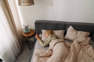 Evde uyuduktan sonra yatakta uzanan genç sarışın kadın cep telefonu kullanıyor.