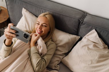 Evde uyuduktan sonra yatakta uzanan genç sarışın kadın cep telefonu kullanıyor.