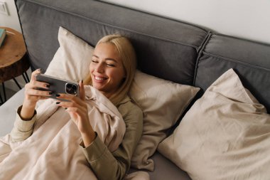 Evde uyuduktan sonra yatakta uzanan genç sarışın kadın cep telefonu kullanıyor.