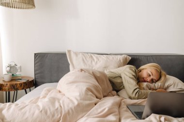 Genç sarışın kadın evde uyuduktan sonra dizüstü bilgisayar kullanıyor.