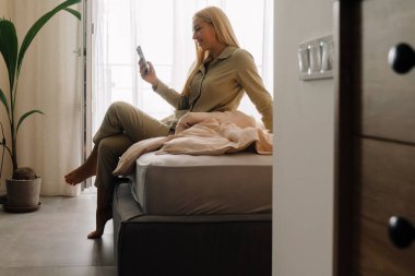 Genç sarışın kadın evde uyuduktan sonra yatakta otururken cep telefonu kullanıyor.