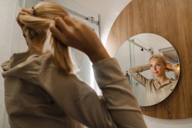 Genç sarışın kadın banyoda saç stili yaparken aynaya bakıyor.