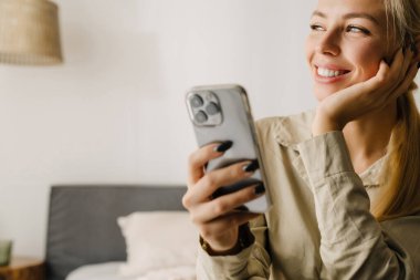 Genç sarışın kadın evde uyuduktan sonra yatakta otururken cep telefonu kullanıyor.