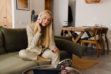 Sarışın kadın makyaj yapıyor ve evdeki koltukta otururken aynaya bakıyor.