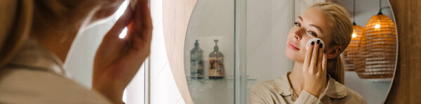 Молодая блондинка, смотрящая в зеркало, когда мыла лицо в ванной