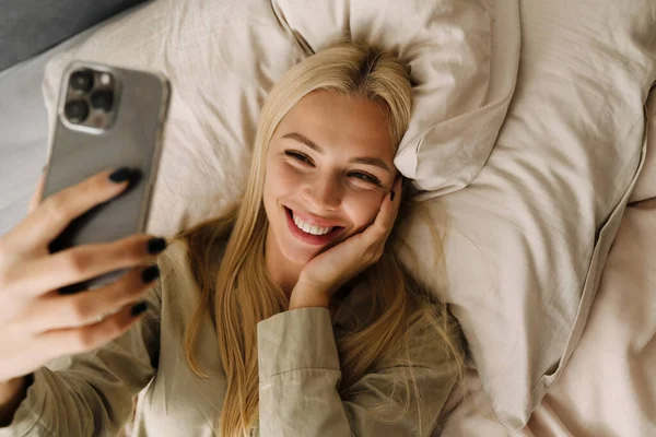 Young blonde woman taking selfie photo while lying in bed after sleep at home