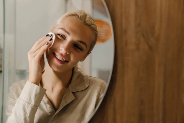 Young blonde woman looking in mirror while cleaning her face at bathroom