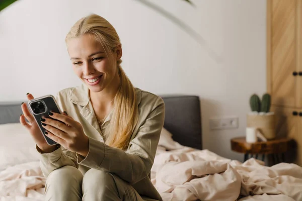 Young blonde woman using mobile phone while sitting on bed after sleep at home