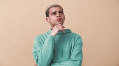 Yoğunlaşmış kıvırcık saçlı, mavi kapüşonlu adam bej rengi stüdyoda düşünüyor.