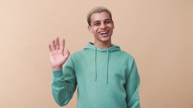 Neşeli kıvırcık saçlı, mavi kapüşonlu adam bej stüdyoda kameraya el sallıyor.
