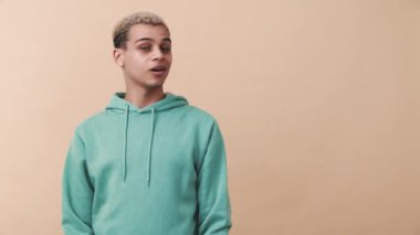 Mavi kapüşonlu neşeli kıvırcık saçlı adam bej stüdyodaki boşluğu işaret ediyor.