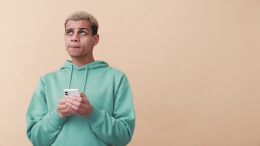 Mavi kapüşonlu neşeli kıvırcık saçlı adam bej stüdyoda telefonla yazı yazıyor.