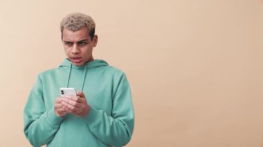 Kıvırcık saçlı, mavi kapüşonlu adam bej stüdyoda telefonla yazı yazıyor.