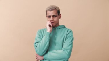 Endişeli kıvırcık saçlı, mavi kapüşonlu adam bej renkli stüdyoya bakıyor.