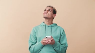 Hoşnut olmayan kıvırcık saçlı, mavi kapüşonlu adam bej stüdyoda telefonla yazı yazıyor.