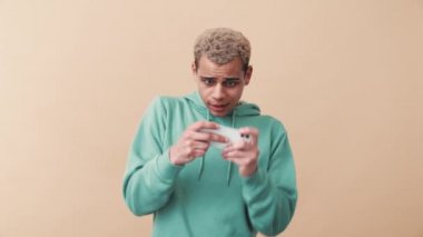 Mavi kapüşonlu mutlu kıvırcık saçlı adam bej stüdyoda telefonla dijital oyun oynuyor.