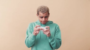 Hevesli kıvırcık saçlı mavi kapüşonlu adam bej stüdyoda telefonla dijital oyun oynuyor.