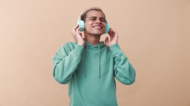 Mavi kapüşonlu, bej renkli stüdyoda kulaklıkla müzik dinleyen, mutlu kıvırcık saçlı bir adam.