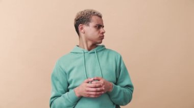 Hoşnut olmayan kıvırcık saçlı mavi kapüşonlu adam bej stüdyoyu işaret ediyor.