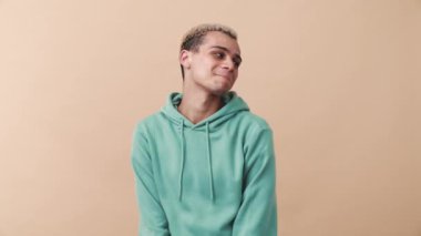 Kafası karışmış kıvırcık saçlı mavi kapüşonlu adam bej stüdyodaki kameraya bakıyor.