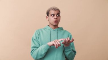Mavi kapüşonlu dalgın kıvırcık saçlı adam bej stüdyoda parmaklarını sayıyor.