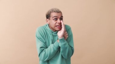 Mavi kapüşonlu yakışıklı kıvırcık saçlı adam sırrını bej stüdyodaki kameraya söylüyor.