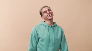 Mavi kapüşonlu neşeli kıvırcık saçlı adam bej stüdyodaki kameraya veda ediyor.