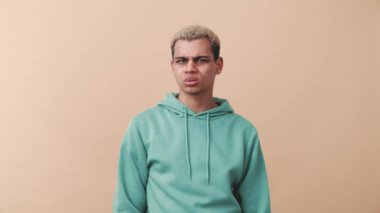 Hoşnut olmayan kıvırcık saçlı mavi kapüşonlu adam bej stüdyodaki kameraya hayır diyor.