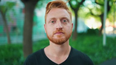 Ciddi kızıl saçlı bir adam parkta dikilirken kameraya bakıyor.